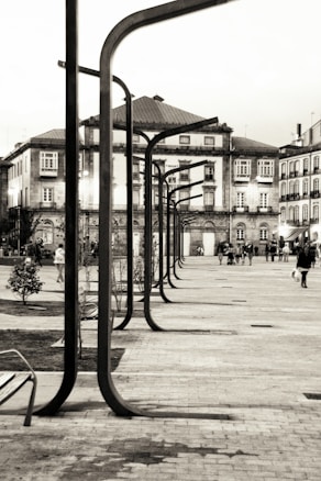 Urban scene featuring a row of modern street lamps leading along a pathway surrounded by historic buildings. People are walking in the area, some in groups and others alone, providing a sense of movement and community. The architecture of the surrounding buildings reflects classical European style with intricate facades and multiple windows.