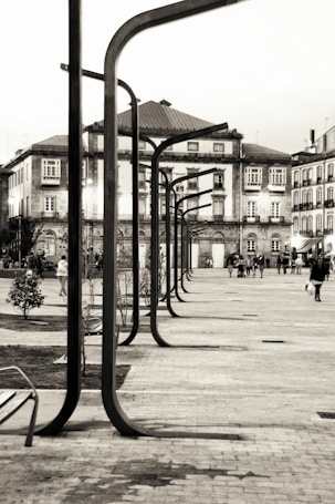 Urban scene featuring a row of modern street lamps leading along a pathway surrounded by historic buildings. People are walking in the area, some in groups and others alone, providing a sense of movement and community. The architecture of the surrounding buildings reflects classical European style with intricate facades and multiple windows.
