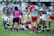 A group of rugby players on a grassy field engaged in a game. Players are wearing distinct team colors, with one team in purple and white stripes and another in red and grey. An official wearing a pink and black uniform is in the center, appearing to be making a call. One player is on the ground holding the ball, while others are gathered closely around.