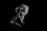 A dancer is captured in profile against a black background, poised in a graceful, crouched position. Flourishes of powder or smoke billow dramatically around their head, creating a dynamic, ethereal effect. Light highlights the dancer's form and the texture of the floating particles.