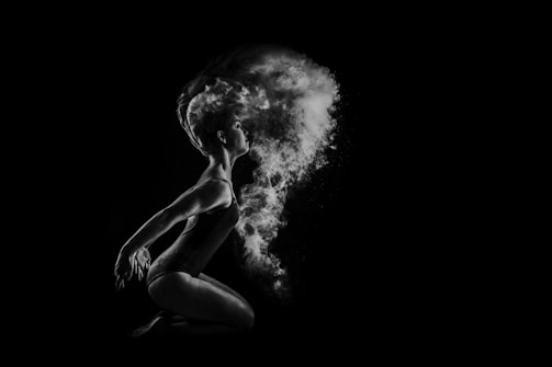 A dancer is captured in profile against a black background, poised in a graceful, crouched position. Flourishes of powder or smoke billow dramatically around their head, creating a dynamic, ethereal effect. Light highlights the dancer's form and the texture of the floating particles.