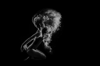 A dancer is captured in profile against a black background, poised in a graceful, crouched position. Flourishes of powder or smoke billow dramatically around their head, creating a dynamic, ethereal effect. Light highlights the dancer's form and the texture of the floating particles.
