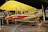 A vintage biplane with a bright yellow top wing and a red lower wing, featuring a circular emblem. The aircraft is displayed indoors with ropes surrounding it, indicating it's part of a museum or exhibition. There are metal supports and propellers, showcasing classic early aviation design. Several people in the background are observing the exhibit.