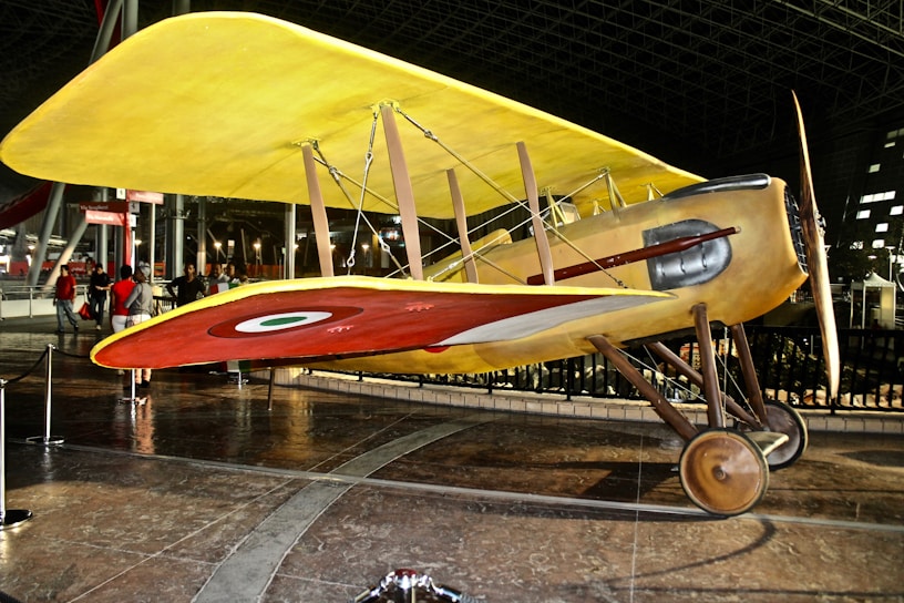 A vintage biplane with a bright yellow top wing and a red lower wing, featuring a circular emblem. The aircraft is displayed indoors with ropes surrounding it, indicating it's part of a museum or exhibition. There are metal supports and propellers, showcasing classic early aviation design. Several people in the background are observing the exhibit.