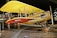 A vintage biplane with a bright yellow top wing and a red lower wing, featuring a circular emblem. The aircraft is displayed indoors with ropes surrounding it, indicating it's part of a museum or exhibition. There are metal supports and propellers, showcasing classic early aviation design. Several people in the background are observing the exhibit.