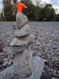 A balanced stack of five large stones is situated in a rocky outdoor area. In the background, a person wearing an orange shirt and running shoes is captured mid-motion, with greenery and trees lining the area.