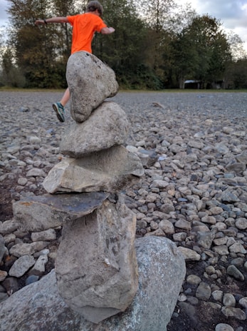 A balanced stack of five large stones is situated in a rocky outdoor area. In the background, a person wearing an orange shirt and running shoes is captured mid-motion, with greenery and trees lining the area.