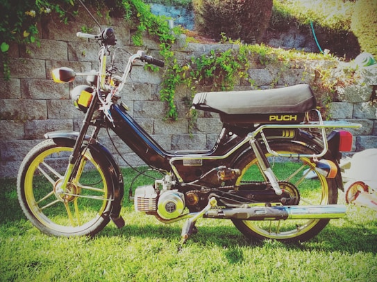 A vintage moped with a black frame and yellow detailing is parked on a grassy lawn. It features classic design elements with visible engine components and a shiny chrome exhaust. The background has a stone wall covered in green ivy, which adds to the nostalgic and serene setting.