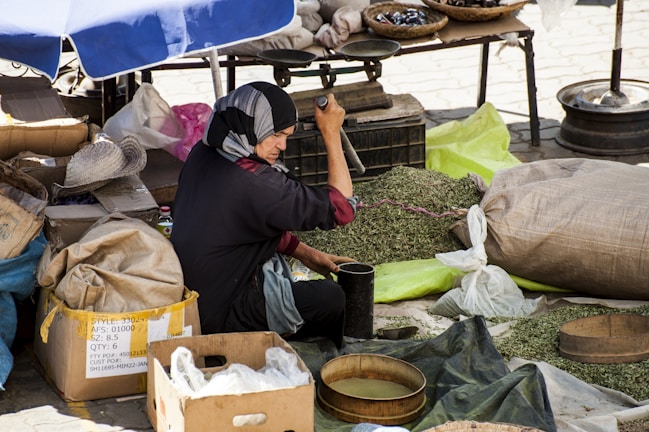 A person wearing a headscarf is seated on the ground, engaged in grinding green leaves with a large mortar and pestle at an outdoor market. Surrounding them are various bags and boxes, some filled with more green leaves. A mix of umbrellas and canopies provide shade, casting shadows over the scene.