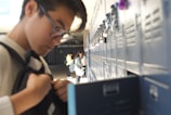 Students are at a row of lockers in a school hallway. One student in the foreground is adjusting their backpack while others in the background access their lockers. The scene is brightly lit with natural light, creating a relaxed atmosphere.