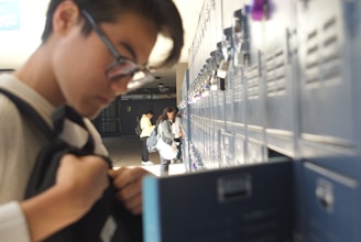 Students are at a row of lockers in a school hallway. One student in the foreground is adjusting their backpack while others in the background access their lockers. The scene is brightly lit with natural light, creating a relaxed atmosphere.