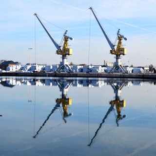 Two large cranes stand on a loading dock, reflected clearly in the calm water of a harbor. The cranes are yellow with grey arms, and the background contains stacks of white materials and several trees. The sky is blue with a few clouds.
