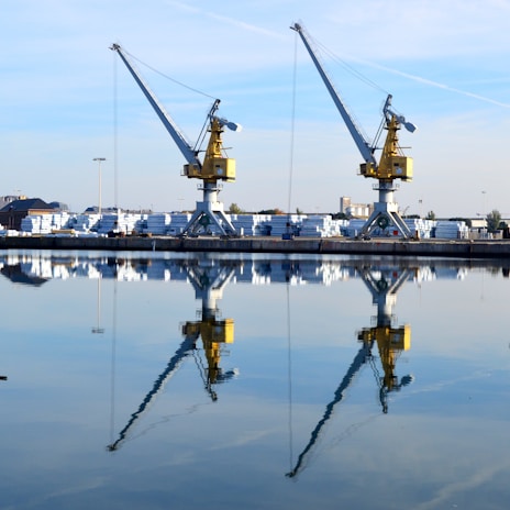 Two large cranes stand on a loading dock, reflected clearly in the calm water of a harbor. The cranes are yellow with grey arms, and the background contains stacks of white materials and several trees. The sky is blue with a few clouds.