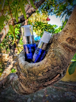 Two bottles of product with blue labeling are nestled in the crook of a tree. The background is a lush outdoor scene with green leaves and branches, creating a natural setting.