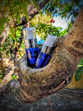 Two bottles of product with blue labeling are nestled in the crook of a tree. The background is a lush outdoor scene with green leaves and branches, creating a natural setting.
