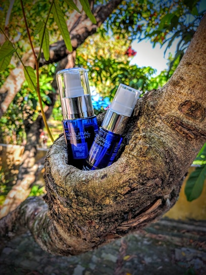 Two bottles of product with blue labeling are nestled in the crook of a tree. The background is a lush outdoor scene with green leaves and branches, creating a natural setting.