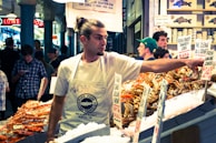 A man with long hair tied back is working at a seafood market, arranging fresh crab and shrimp on ice. He wears a white apron with a logo for the Pure Food Fish Market. Behind him, several other people are visible, some browsing or engaged in conversation. Various signs with seafood prices are prominently displayed.