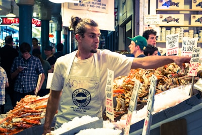 A man with long hair tied back is working at a seafood market, arranging fresh crab and shrimp on ice. He wears a white apron with a logo for the Pure Food Fish Market. Behind him, several other people are visible, some browsing or engaged in conversation. Various signs with seafood prices are prominently displayed.