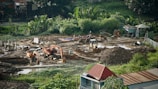 A construction site surrounded by lush greenery, featuring heavy machinery such as excavators and bulldozers working on moving soil. There are several piles of dirt and concrete pillars scattered throughout the site. Workers in hard hats are visible, indicating ongoing labor. The background includes small structures and dense vegetation.