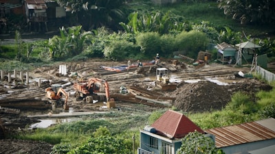 A construction site surrounded by lush greenery, featuring heavy machinery such as excavators and bulldozers working on moving soil. There are several piles of dirt and concrete pillars scattered throughout the site. Workers in hard hats are visible, indicating ongoing labor. The background includes small structures and dense vegetation.
