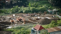 A construction site surrounded by lush greenery, featuring heavy machinery such as excavators and bulldozers working on moving soil. There are several piles of dirt and concrete pillars scattered throughout the site. Workers in hard hats are visible, indicating ongoing labor. The background includes small structures and dense vegetation.