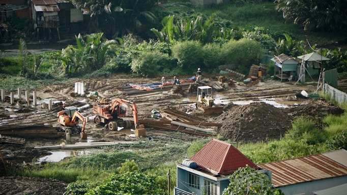 A construction site surrounded by lush greenery, featuring heavy machinery such as excavators and bulldozers working on moving soil. There are several piles of dirt and concrete pillars scattered throughout the site. Workers in hard hats are visible, indicating ongoing labor. The background includes small structures and dense vegetation.