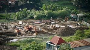 A construction site surrounded by lush greenery, featuring heavy machinery such as excavators and bulldozers working on moving soil. There are several piles of dirt and concrete pillars scattered throughout the site. Workers in hard hats are visible, indicating ongoing labor. The background includes small structures and dense vegetation.