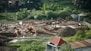 A construction site surrounded by lush greenery, featuring heavy machinery such as excavators and bulldozers working on moving soil. There are several piles of dirt and concrete pillars scattered throughout the site. Workers in hard hats are visible, indicating ongoing labor. The background includes small structures and dense vegetation.