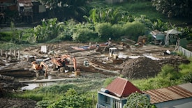 A construction site surrounded by lush greenery, featuring heavy machinery such as excavators and bulldozers working on moving soil. There are several piles of dirt and concrete pillars scattered throughout the site. Workers in hard hats are visible, indicating ongoing labor. The background includes small structures and dense vegetation.