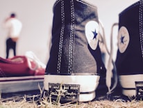 A pair of black and white Converse All Star sneakers is prominently displayed in the foreground. One shoe is in sharp focus while the other is slightly to its side. The background features a blurry figure of a person dressed casually. The ground is covered with grass and gives a slightly rugged feel.