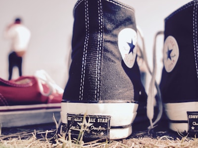 A pair of black and white Converse All Star sneakers is prominently displayed in the foreground. One shoe is in sharp focus while the other is slightly to its side. The background features a blurry figure of a person dressed casually. The ground is covered with grass and gives a slightly rugged feel.