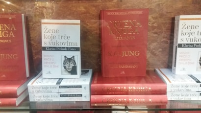 Several books are displayed on a shelf. The prominent titles include a book with a red cover and gold lettering, along with several copies of a white book with a black and white illustration and text in a different language.
