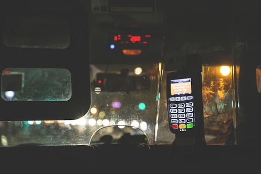 An interior view of a vehicle at night, featuring a bright digital fare meter with glowing lights. The windshield reflects multiple blurred city lights in the background, creating a sense of movement and urban life.