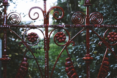 An ornate metal fence with intricate scrollwork and floral designs stands in front of a lush green background. The fence is painted a deep, rusty red color and has graffiti or writing in white scattered across various parts. The forested area behind the fence is slightly blurred, creating depth.