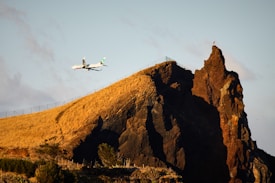 A commercial airplane is flying close to a rugged, rocky mountain with a mix of light and dark stone patterns. The mountain has a steep slope covered in dry, yellowish grass. A fence runs along the ridge. The sky is clear with a few clouds, and one bird is perched on top of the mountain peak.