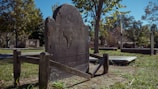 A weathered, dark gravestone stands in an old cemetery, surrounded by green grass and scattered leaves. The gravestone has engravings on it and is supported by a wooden frame. In the background, there are more gravestones and trees with dense green foliage under a clear blue sky.