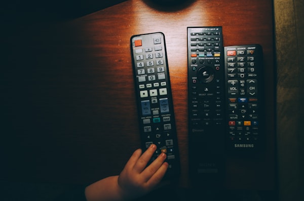 a child's hand reaching for a remote control, next to two others, on a dark wood table under a lamp