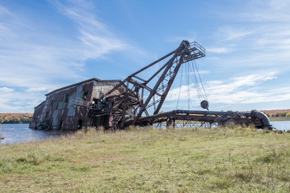 A large, rusted industrial structure or dredge is partially submerged in water, with an extensive metal framework and bucket ladder visible. It is situated near a grassy shoreline under a clear blue sky. In the background, autumn-colored trees line the horizon.
