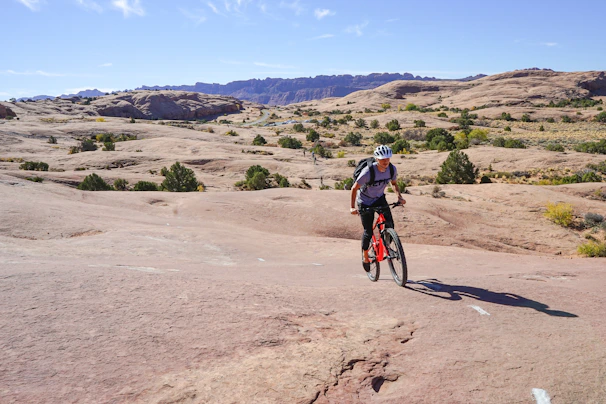 Mountain biker launching off a rocky trail with dust flying in the air under a cloudy sky.