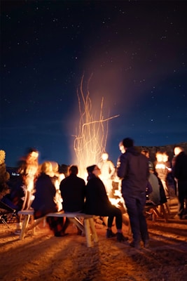 A group of people are gathered around a glowing campfire at night, under a starry sky. They are sitting on logs and benches, some standing, all facing the fire. The scene is illuminated by the warm light of the fire, with sparks and smoke rising into the night.