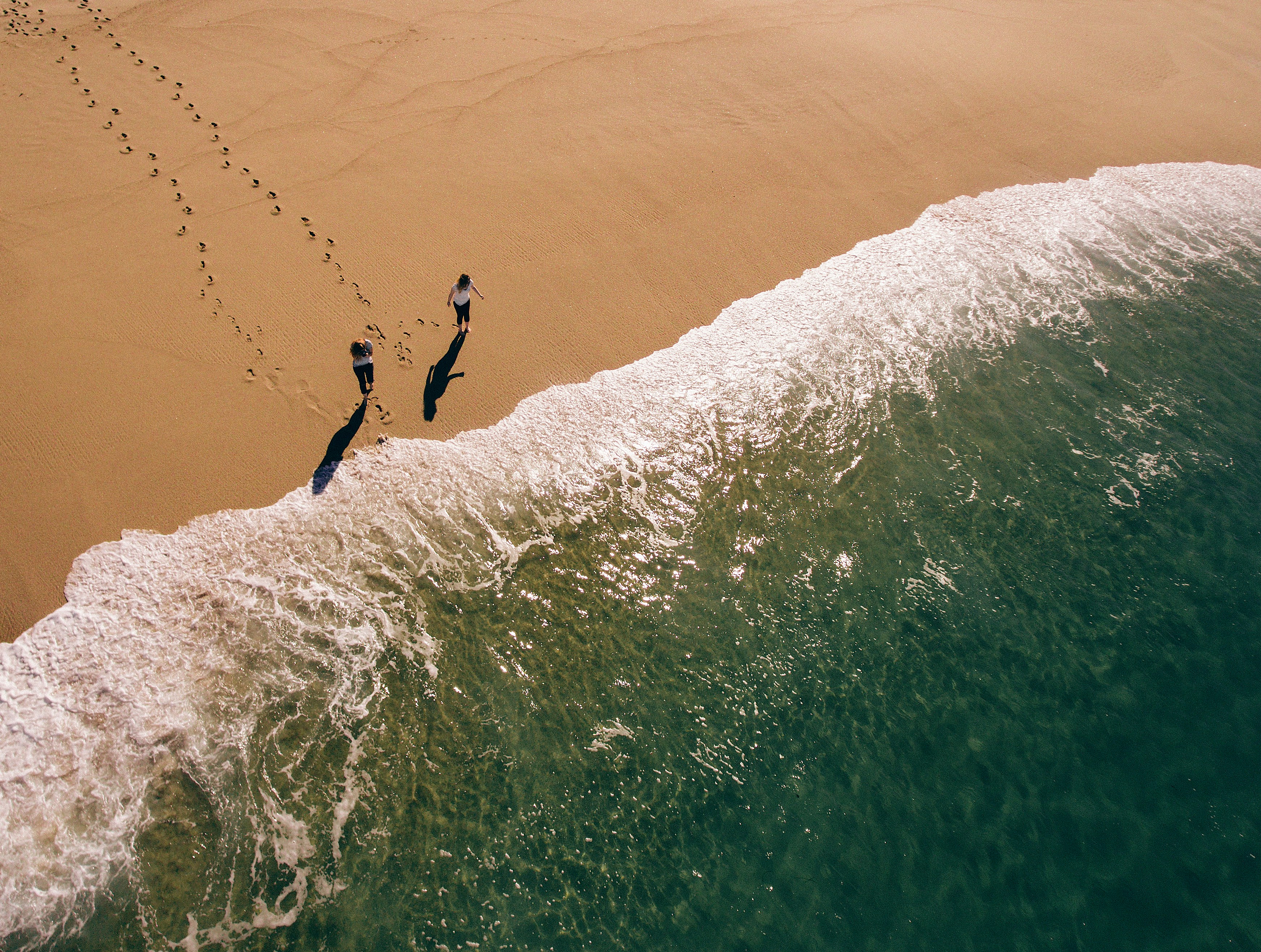 2 people walking in the sand towards the beach waves leaving footsteps