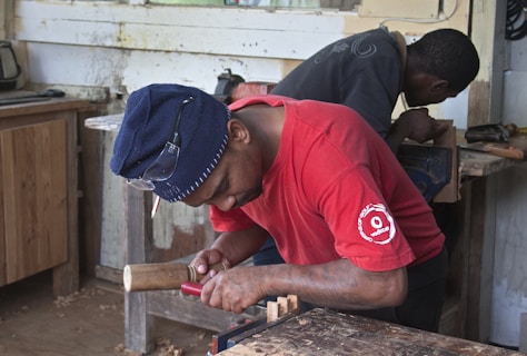 Two individuals are engaged in woodworking in a workshop. The person in the foreground is intently focusing on carving wood with a chisel and mallet, while the other person in the background is also working on a wooden piece. Various tools and wooden materials are scattered around the workshop.
