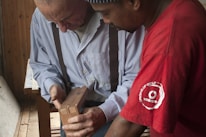 Two men are closely examining a piece of wooden work in a woodworking shop. One man is wearing a striped shirt with suspenders, while the other is in a red t-shirt. They appear to be discussing or teaching something related to the woodwork.