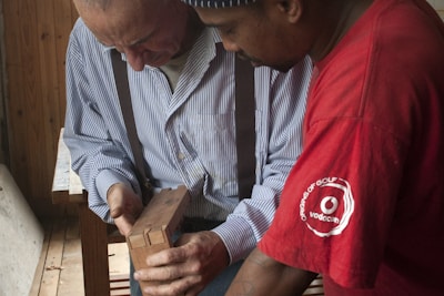 Two men are closely examining a piece of wooden work in a woodworking shop. One man is wearing a striped shirt with suspenders, while the other is in a red t-shirt. They appear to be discussing or teaching something related to the woodwork.