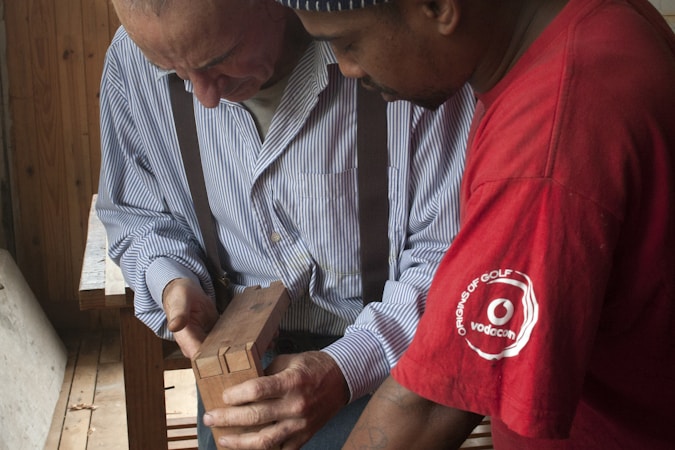 Two men are closely examining a piece of wooden work in a woodworking shop. One man is wearing a striped shirt with suspenders, while the other is in a red t-shirt. They appear to be discussing or teaching something related to the woodwork.