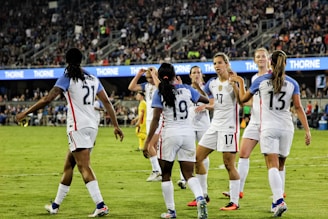 A group of female soccer players in white jerseys, with blue sleeves and red accents, are celebrating on a green field. They are high-fiving and appear to be in good spirits. The background shows a stadium filled with spectators, creating an energetic atmosphere.