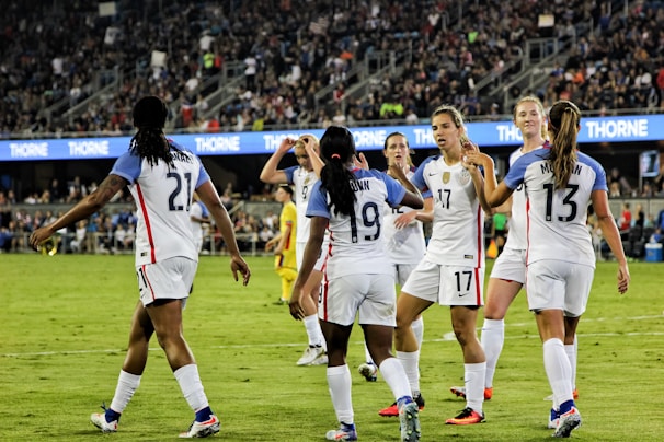 A group of female soccer players in white jerseys, with blue sleeves and red accents, are celebrating on a green field. They are high-fiving and appear to be in good spirits. The background shows a stadium filled with spectators, creating an energetic atmosphere.