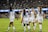 A group of female soccer players in white jerseys, with blue sleeves and red accents, are celebrating on a green field. They are high-fiving and appear to be in good spirits. The background shows a stadium filled with spectators, creating an energetic atmosphere.