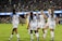 A group of female soccer players in white jerseys, with blue sleeves and red accents, are celebrating on a green field. They are high-fiving and appear to be in good spirits. The background shows a stadium filled with spectators, creating an energetic atmosphere.