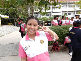 A group of people wearing white and pink uniforms are gathered outdoors in what appears to be a school setting. A person in the foreground is smiling and making a peace sign. The background includes more people, trees, and a building with windows.
